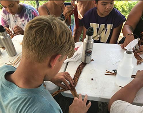 A young boy sits at a table surrounded by other children, crafting a cedar headband